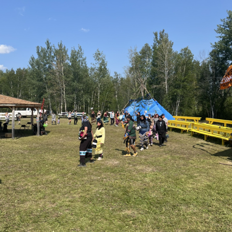 Photo of a group of people in a grass field with a teepee and bleachers in the background.